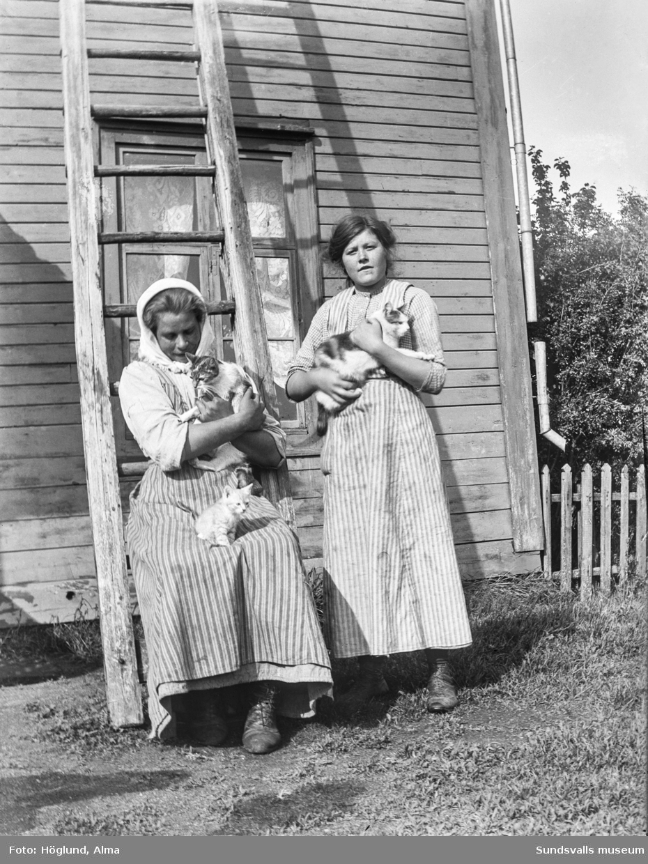 Två unga kvinnor med katter i famnen invid en stege som står lutad mot en husvägg.
