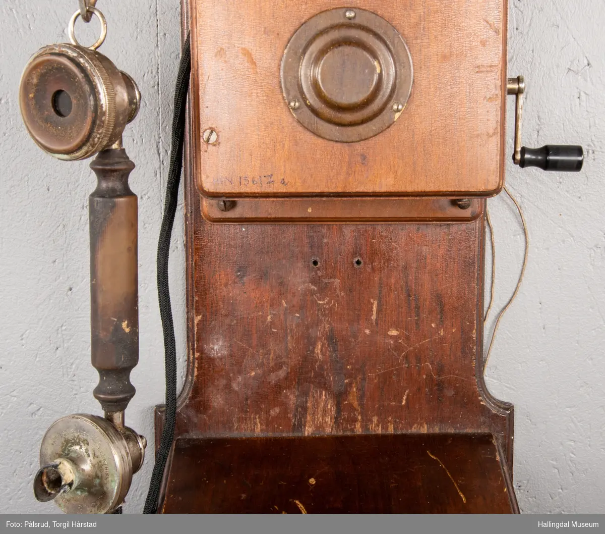 En veggtelefon i tre med to ringeklokker i blank metall øverst på boks. Svart leding koblet til telefonrør og sveiv med svart plasthåndtak på motsatt side. Flere ledninger på baksiden. Nederst en boks i tre for oppbevaring av to batterier produsert av Norsk Akkumulator Co. A/S - påskrevet dato 1953. Telefonen har tilhørende skriv med regler for telefonbruk for Tunhøvd - Næstelefon, med dato 1918. 