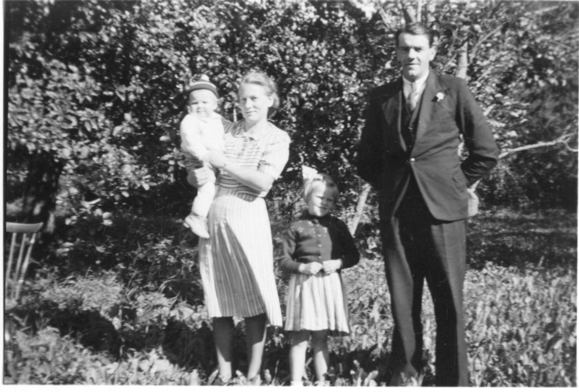 En man, en kvinna och två barn. Kvinnan håller den minsta i famnen. De står troligen i en trädgård.