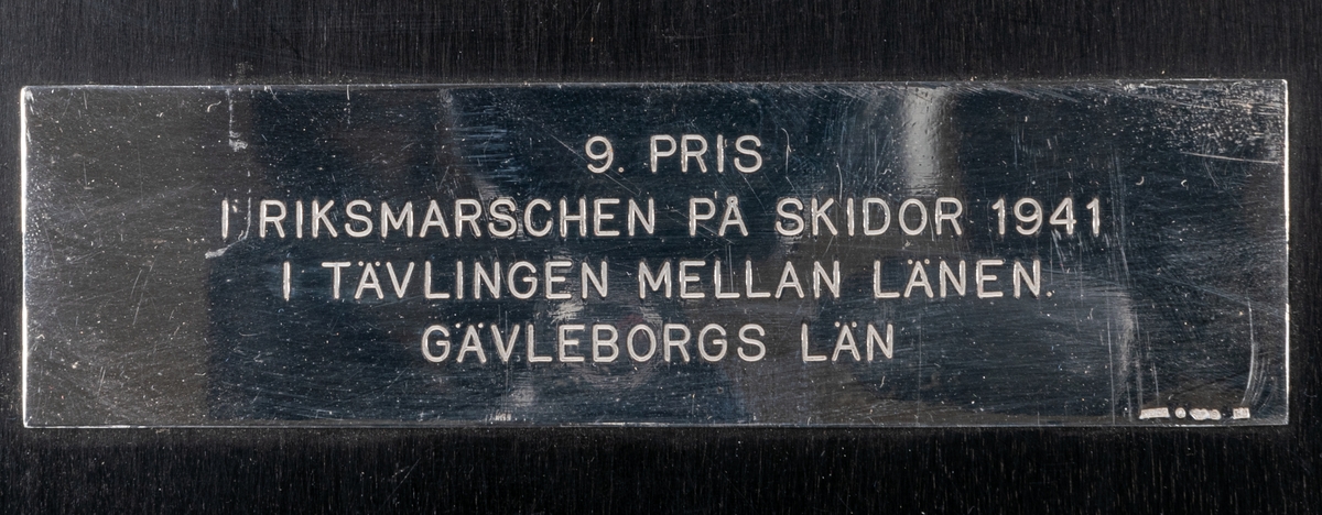 Plakett, brons. "Fädrens spår" monterad på svart trätavla. Silverskylt: "9 Pris i Riksmarschen på skidor 1941 i tävlingen mellan länen. Gefleborgs län."
