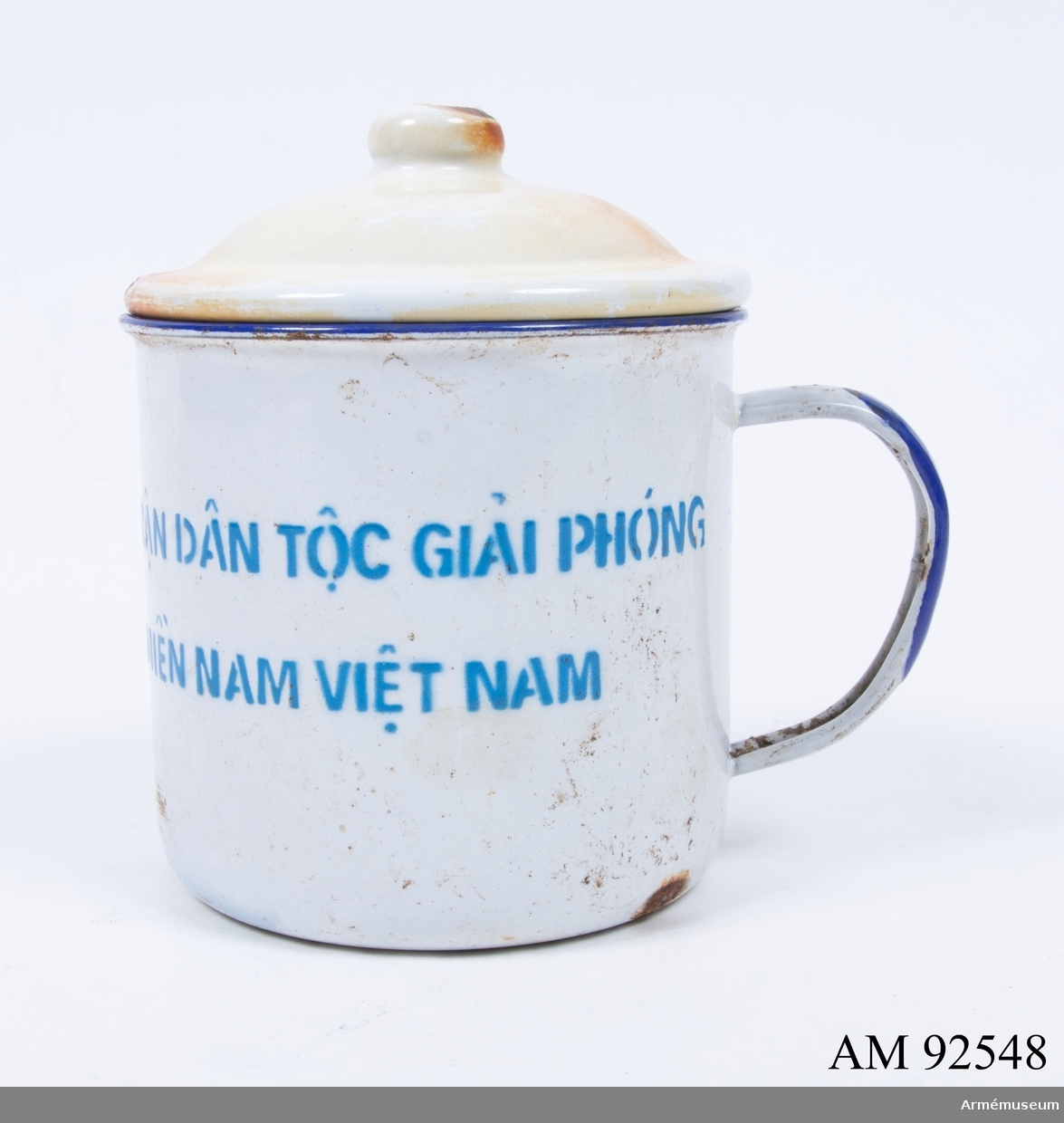 Mugg med lock. Emaljerad i vitt och dekorerad med flagga för ”Front populaire pour la libération du Sud du Vietnam” (FNL, eller ”Viet Cong”), samt med vietnamesisk text (i översättning) ”Nationella Frihetsfronten, Syd Vietnam”. Botten med årtalet ”1968”. Vietnam, Vietnamkriget.

På muggen står:
MAT TRAN DAN TOC GIAI PHONG/MIEN NAM VIET NAM