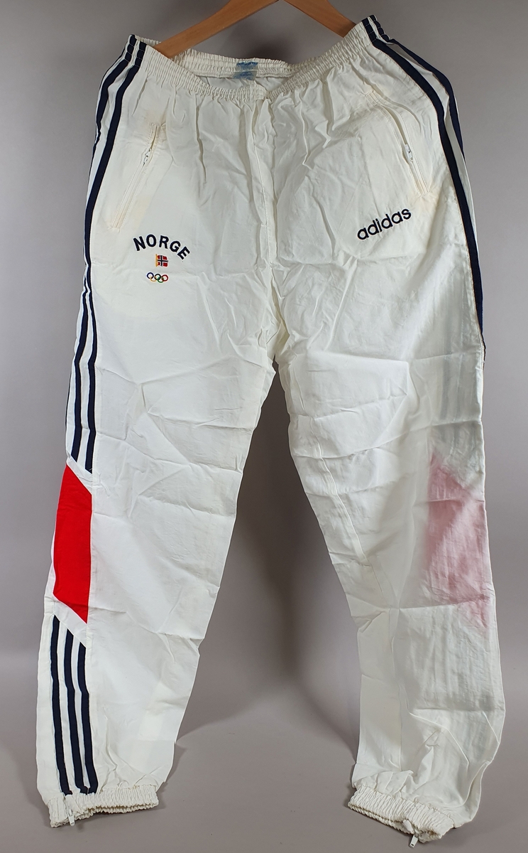 To hvite treningsbukser med striper i rødt og blått på ytterside av hvert ben. På høyre hofte er det skrevet NORGE, med de olympiske ringer og det norske flagg. På venstre hofte logo for Adidas.