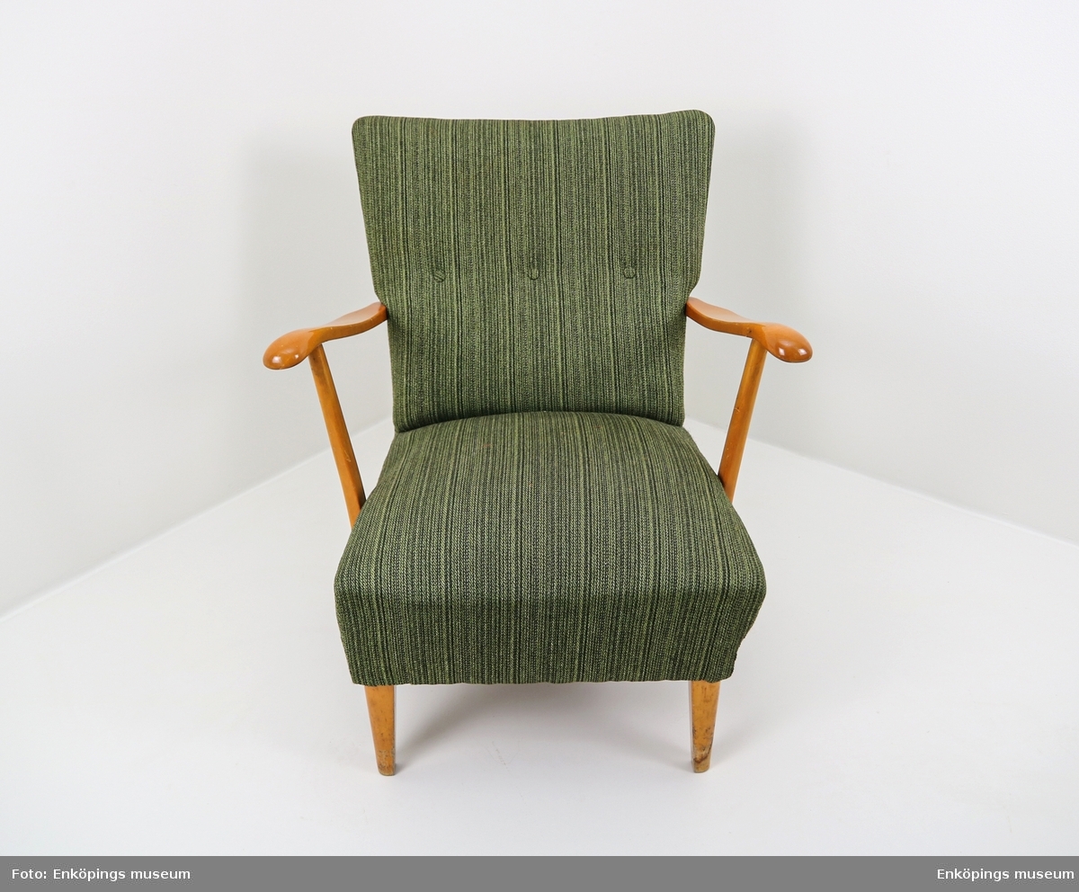 Grön fåtölj från 1950-talet. Ljust träslag i armstöd och ben. Sits och ryggstöd klädda med vävt tyg. På stolsryggens framsida finns tre textilklädda knappar.