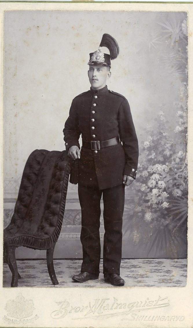 Kabinettsfotografi av en ung man i militär uniform. Han står med höger hand vilande på en stolsrygg.