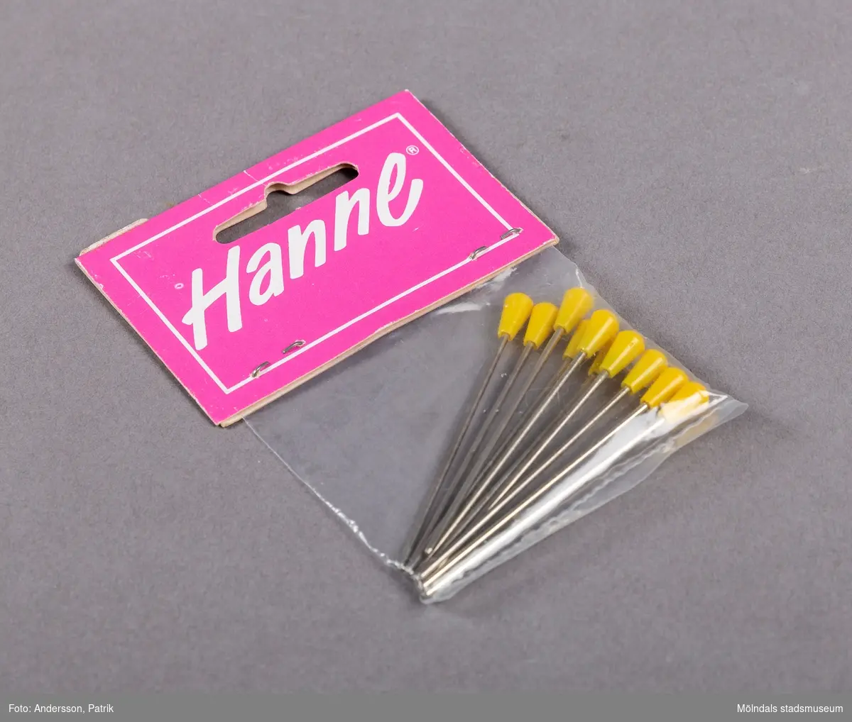 En mindre plastpåse innehållande nålar med gul ände av plast. Etiketten som är rosa, innehåller företagets namn skrivet i vit text, inom en vit ram.
Etiketten är fasthäftad på påsen med två häftstift.