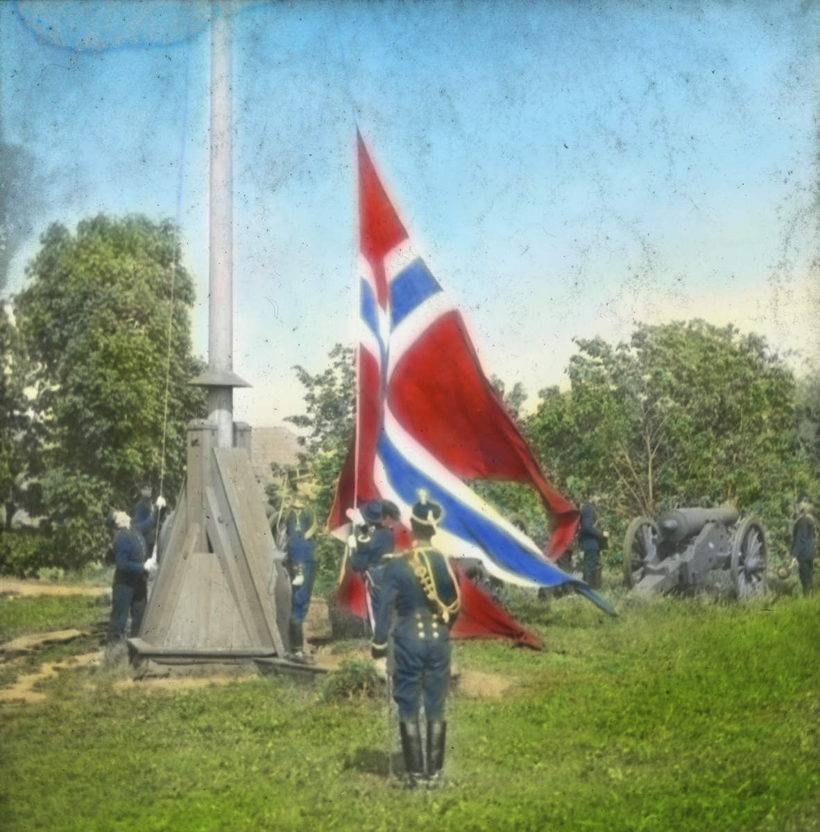 Håndkolorert dias. To offiserer overvåker heisingen av det norske flagget på Akershus festning 9. juni 1905. To soldater heiser flagget, mens to står oppstilt ved en kanon.