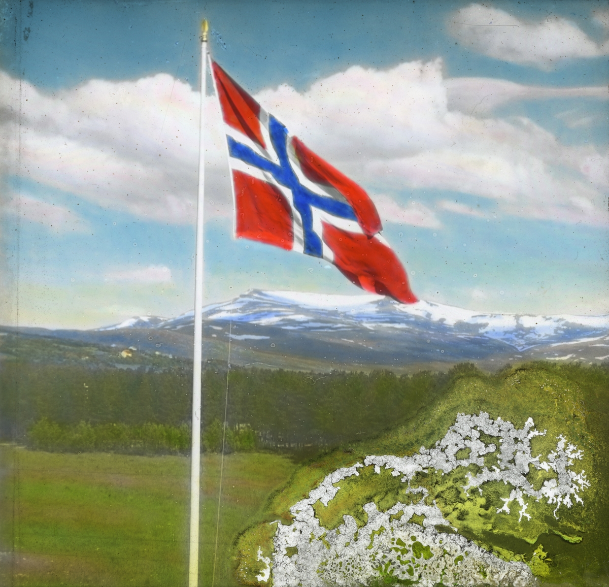 Håndkolorert dias. Et norsk flagg vaier i vinden. I bakgrunnen sees grønne åser og snødekte fjell.