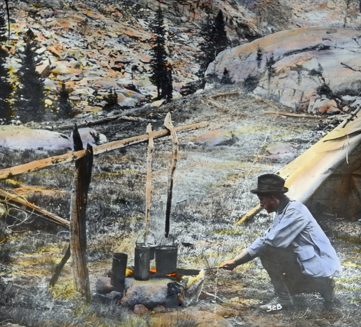 Håndkolorert dias. En mann passer bålet, der to kjeler koker. Bak skimtes delene av et telt og et steinete fjellandskap. I boken "En emigrants ungdomserindringer" forteller fotografen Anders Beer Wilse om sitt arbeid i U.S.A.  