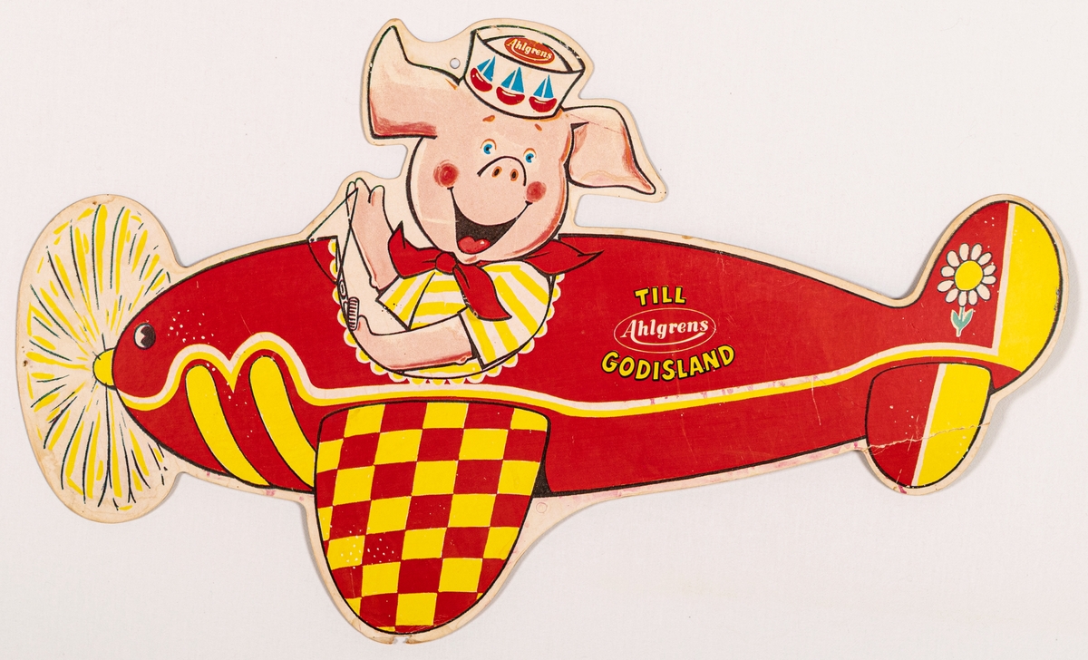 Reklamskylt av papp, flerfärgstryck, för Ahlgrens godis. Tryck på båda sidor. En röd flygmaskin med en gris som pilot.