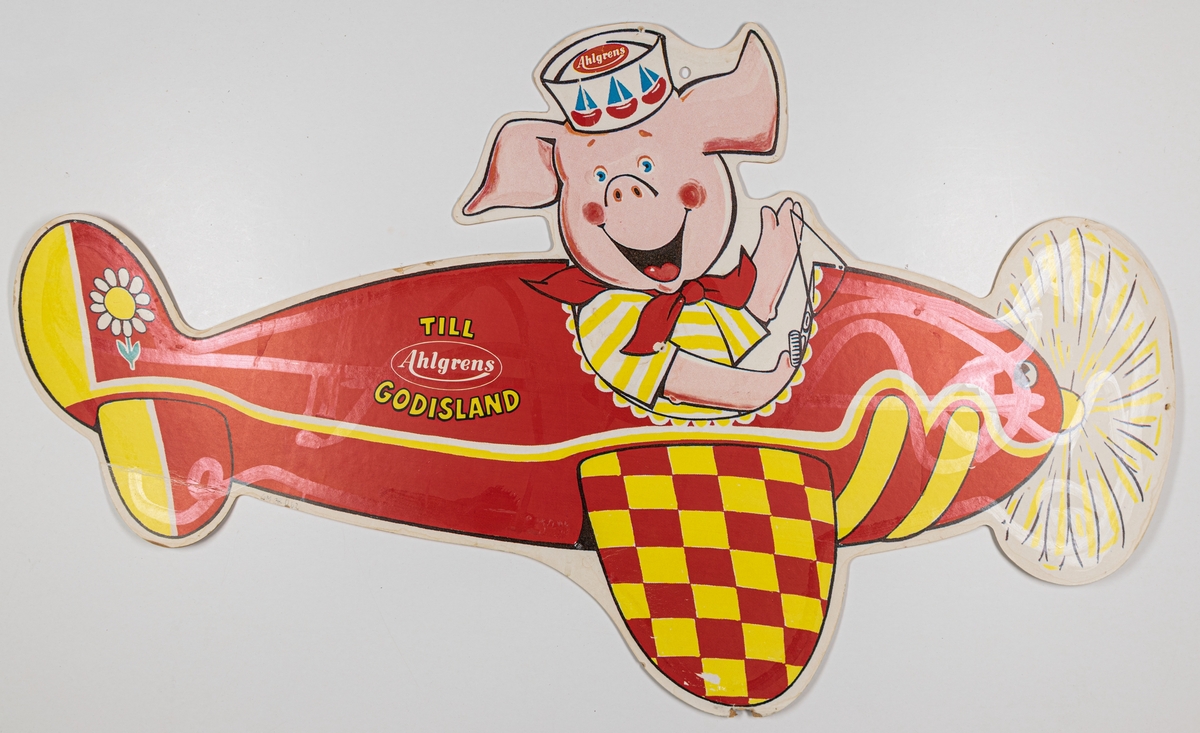 Reklamskylt av papp, flerfärgstryck, för Ahlgrens godis. Tryck på båda sidor. En röd flygmaskin med en gris som pilot.