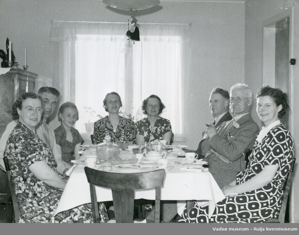 Mennesker samlet rundt spisestuebordet sommeren 1953. Onkel Petter fra Amerika og Harald fra Oslo er på besøk. Heksefigur dingler i taklampa. Vadsø