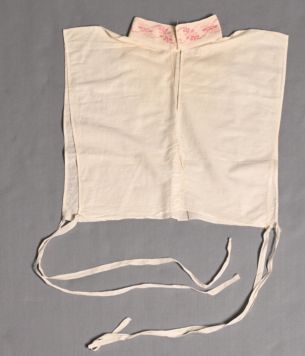 Isättning till grönländsk festdräkt för kvinnor.
Löst skjortbröst av vitt bomullstyg med liten ståkrage med rosa-blekrosa korsstygnsbroderi på aidaväv. Knyts med bomullsband i båda sidorna.