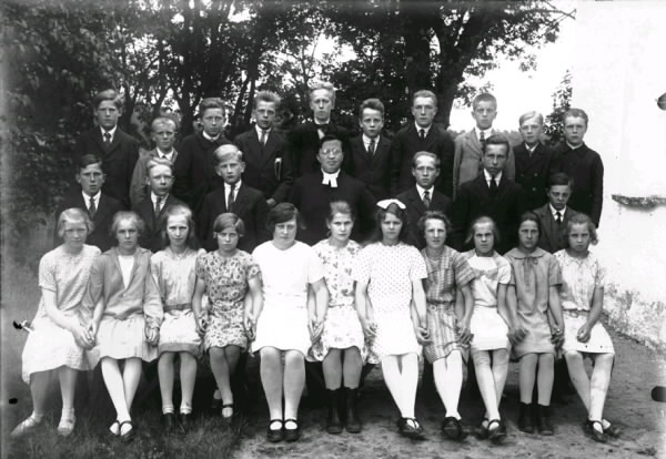 1928-års konfirmander med prästen Johan Hultskog i mitten. Pojkarna har kostym men flickorna har inte någon enhetlig konfirmationsklädsel utan olika mönstrade klänningar och de håller varandra i handen.