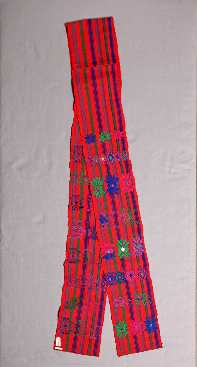 Brett vävt bälte från Santa Maria de Jesus, Guatemala. Hör till kvinnodräkten. 
Rödrandig panama med inplockade mönster i grönt, blått, och två lila nyanser.