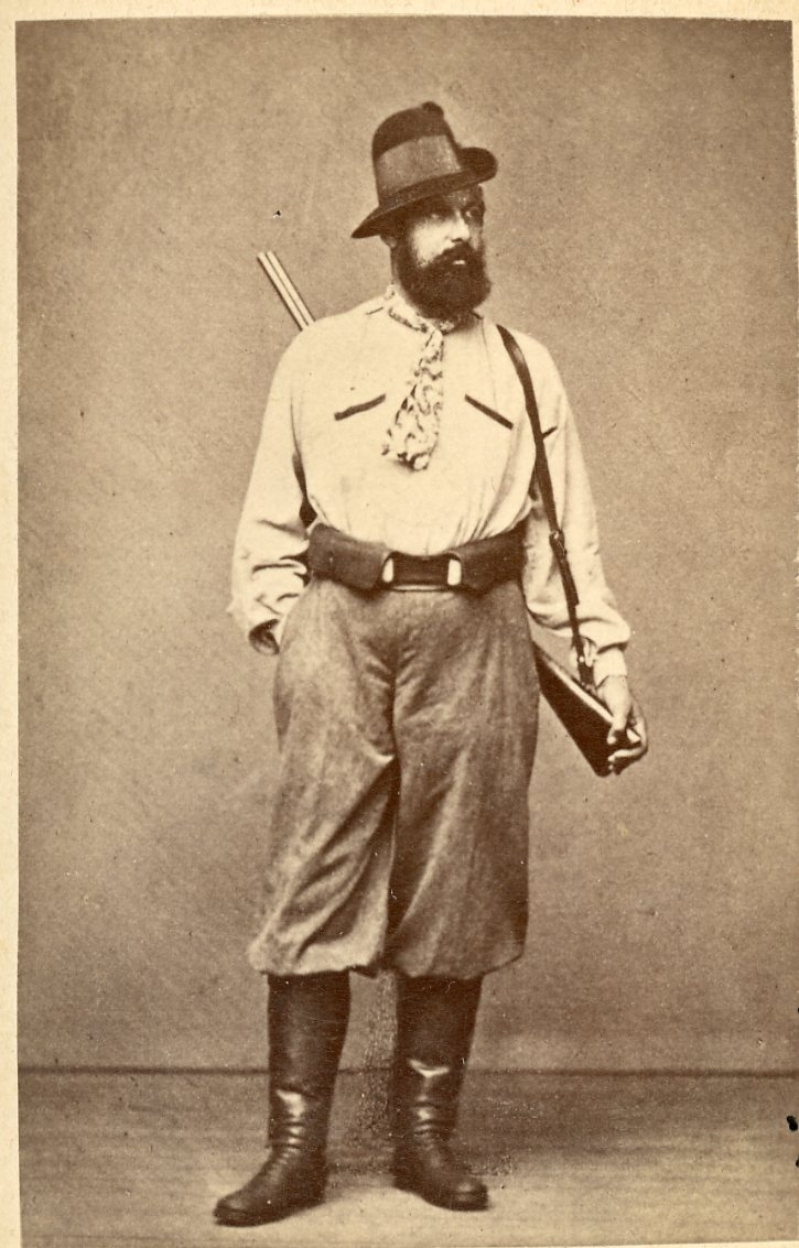 Kabinettsfotografi av en man klädd för jakt, med hatt och scarf runt halsen och ett gevär hängande över axeln.