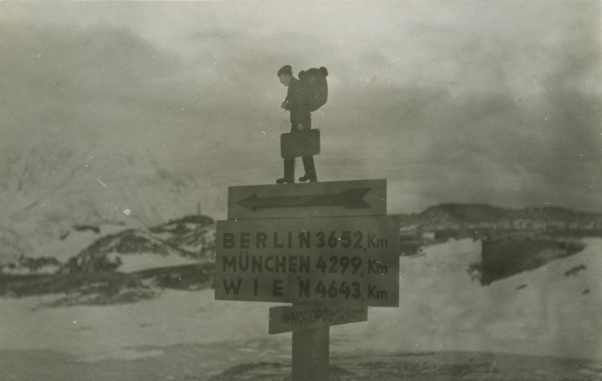 På bildets bakside står det håndskrevet "Wegweiser in Hammerfest", veiviser i Hammerfest. Fotografiet viser en veiviser i vinterlandskap. Skiltene er festet på en firkantet stolpe. Øverst er det festet en utskåren figur av en soldat med stor ryggsekk og koffert. Under den peker en pil til venstre. Det største skiltet informerer om avstandene til Berlin, München og Wien. Nederst viser et skilt veien til "Nordpol 10 km".