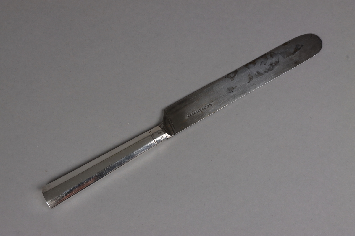 Bordskniv av stål med facetterat skaft av silver. Vid fäste för knivblad dubbla linjer runt om. Skaft med rakt avslut.
