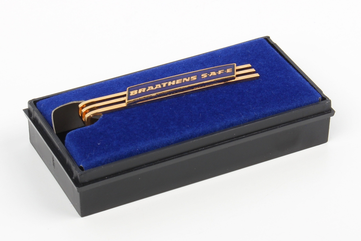 Gullfarget slipsnål med Braathens-logo i blå emalje. Festet på et stykke blå kartong, ligger i lite etui av plast med svart bunn og gjennomsiktig lokk.