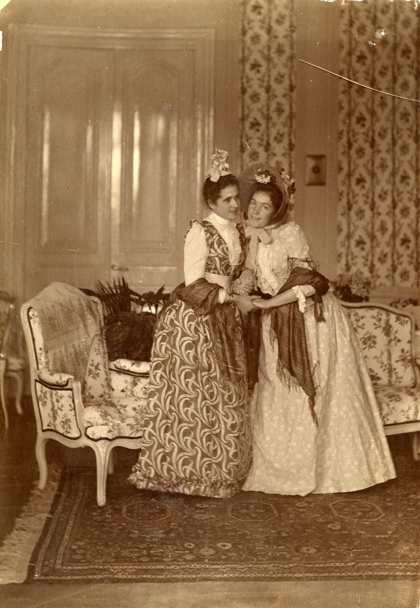To kvinner i pene kjoler fotografert innendørs i en salong. Teppe på gulvet, møbler og en dør i bakgrunnen. Trolig er den ene av kvinnene Sandra Droucker.