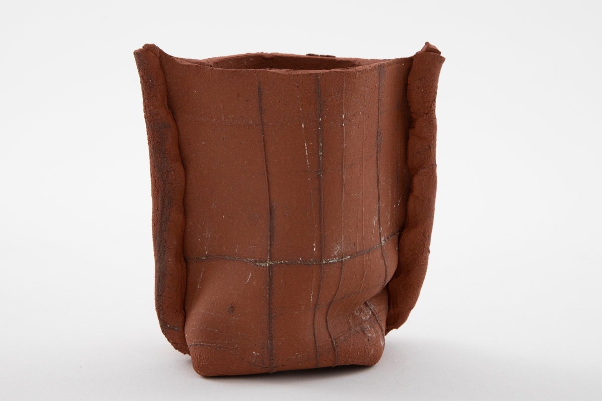 Matt rødbrun vase i steingods med grunnform som en spiss oval og rette vegger som er klemt litt inn nederst. Leireplatene overlapper hverandre på sidene og danner en hanklignende kant. Tekstur i overflaten. Åpen munning.