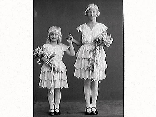 Syskonbild av två flickor i sommarskrud med blomdiadem, volangkjolar och blombuketter. Kamrer Bramstång beställde bilderna, sannolikt barnens far.