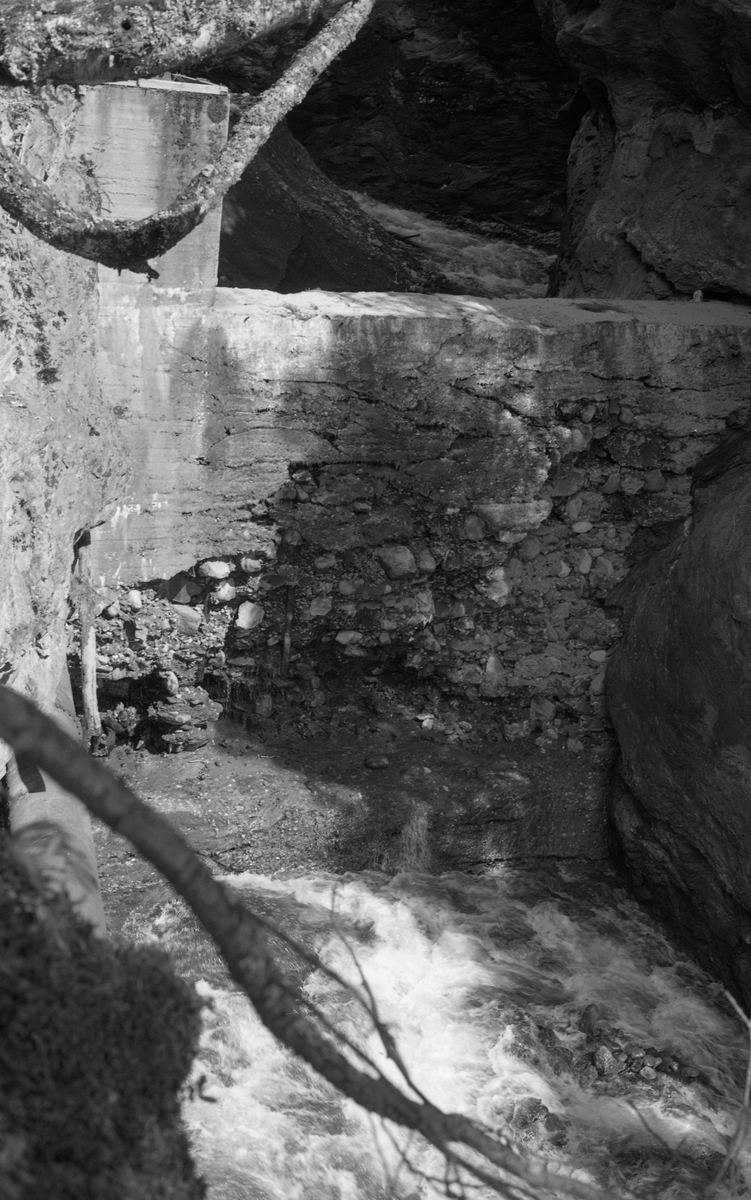 Kraftverksdammen i elva Tromsa med den øvre delen av turbinrøret som førte vann til Tromsa mølles kraftverk på Fåvang i Ringebu kommune i Gudbrandsdalen. Denne delen av turbinrøret var sammensatt støpejernelementer som var skrudd sammen, ende mot ende, og som da dette fotografiet ble tatt var intakt etter flommen i 1958, som førte til brudd i en nedenforliggende del av røret, som var lagd av tre (se SJF.1994-01954). Dammen som magasinerte vann til kraftverket var plassert på et sted der elva gikk gjennom ei trang bergkløft i et elveparti med betydelig fall. Den var støpt av betong, men fotografiet viser at den var kraftig forvitret, antakelig fordi sementen som var brukt var av dårlig kvalitet og fordi man hadde drøyd med atskillig grov stein. Da dette fotografiet ble tatt fosset det fram vann under den nedre delen av dammen.

Hvilke interesser Glomma fellesfløtingsforening, som har eid dette fotografiet, kan ha hatt i forbindelse med skadeflommen i dette vassdraget i 1958, er foreløpig ukjent. Topografien i Tromsas elveløp var meget kupert, og fra gammelt av ble det ikke fløtet tømmer der i det hele tatt. I 1958 var det imidlertid innmeldt 28 325 tømmerstokker til fløting i denne elva. De første stokkene ble utislått 28. mai, like etter flomkatastrofen. Med unntak av 169 stokker skal dette tømmeret ha nådd ned til Gudbrandsdalslågen.