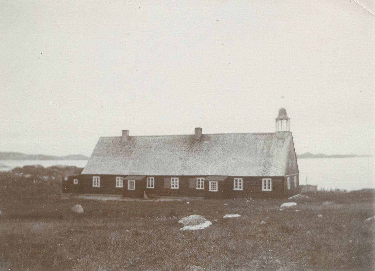 Fotografi från expedition till Grönland. Motiv av träkyrka belägen invid havet. Kyrkan är avlång med nio fönster på ena långsidan och två skorstenar på taket. På taket längst fram sitter ett litet kyrktorn.