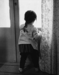 Liten flicka, med ryggen mot fotografen, tittar ut genom ett