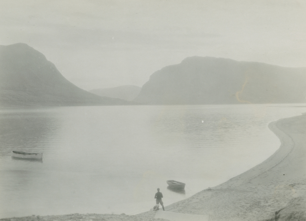 Fotografi från expedition till Grönland. Motiv av Tyrolerfjorden på östra Grönland. På bilden syns en man som står på en strand, med fjorden framför sig och stora berg i bakgrunden.