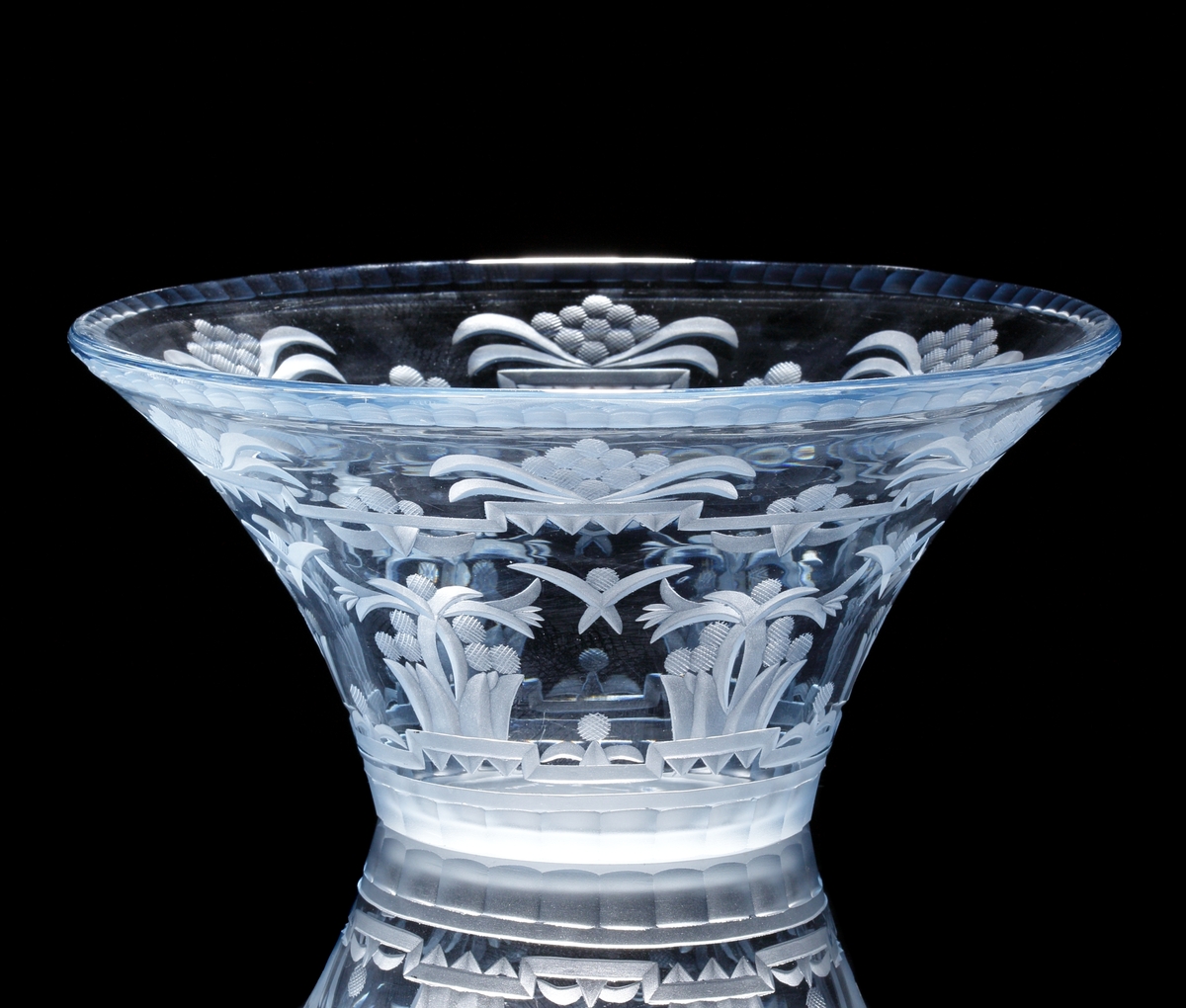 Formgiven av Edward Hald. Ljusblå trattformad skål med fasetterad mynningskant och slipat mönster i form av stiliserade
blomkorgar. Stående fasetter vid skålens nedre kant.