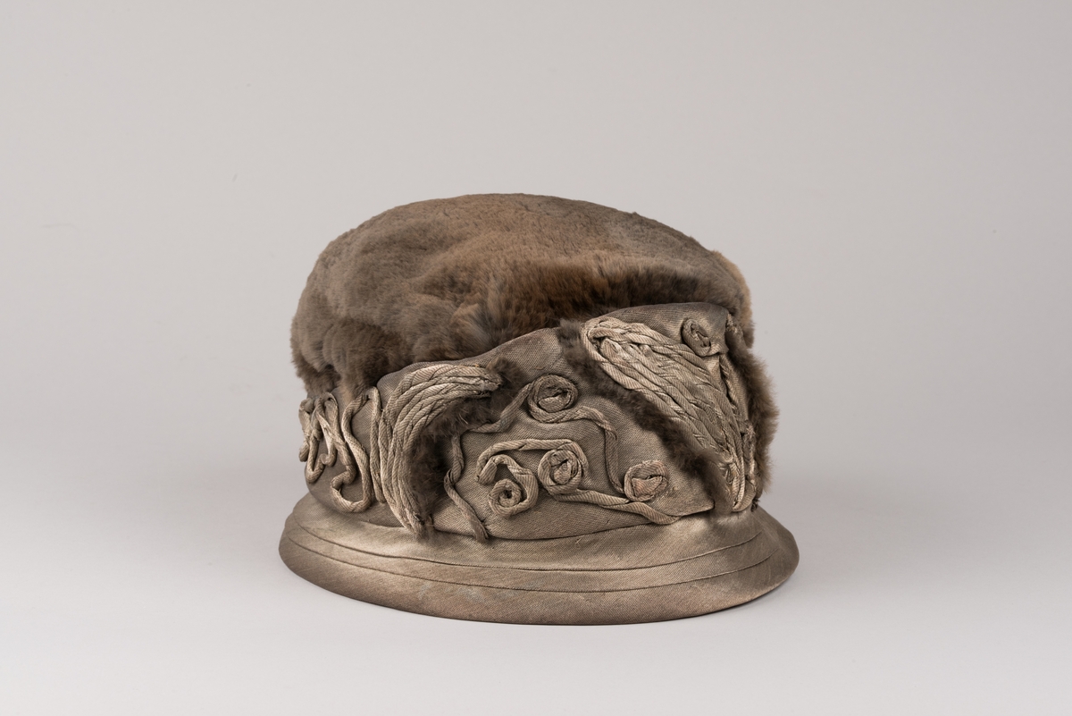 Hatt, modell med brun päls på kullen och grått tyg av metalltråd. Hatten har ett litet styvt brätte och en stärkt kant som är bredare i framkant. Dekorationer av tvinnat tyg och päls.