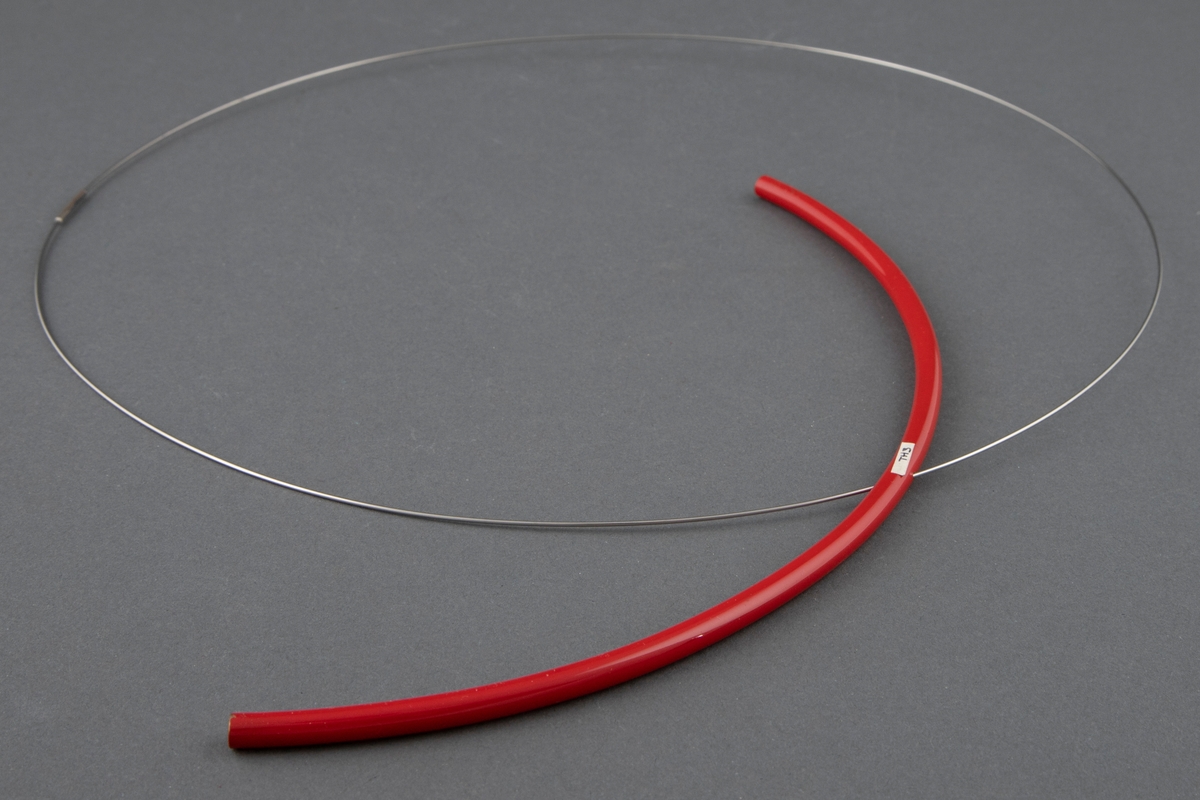 Halssmykke bestående av en tynn stålring der det er tredd på en halvsirkelformet, rødlakkert messingstav.