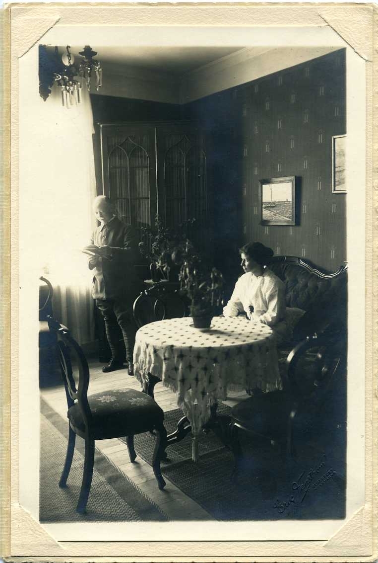 En kvinna sitter i en soffa vid ett bord. En man står vid ljuset från ett fönster och tittar i en bok.