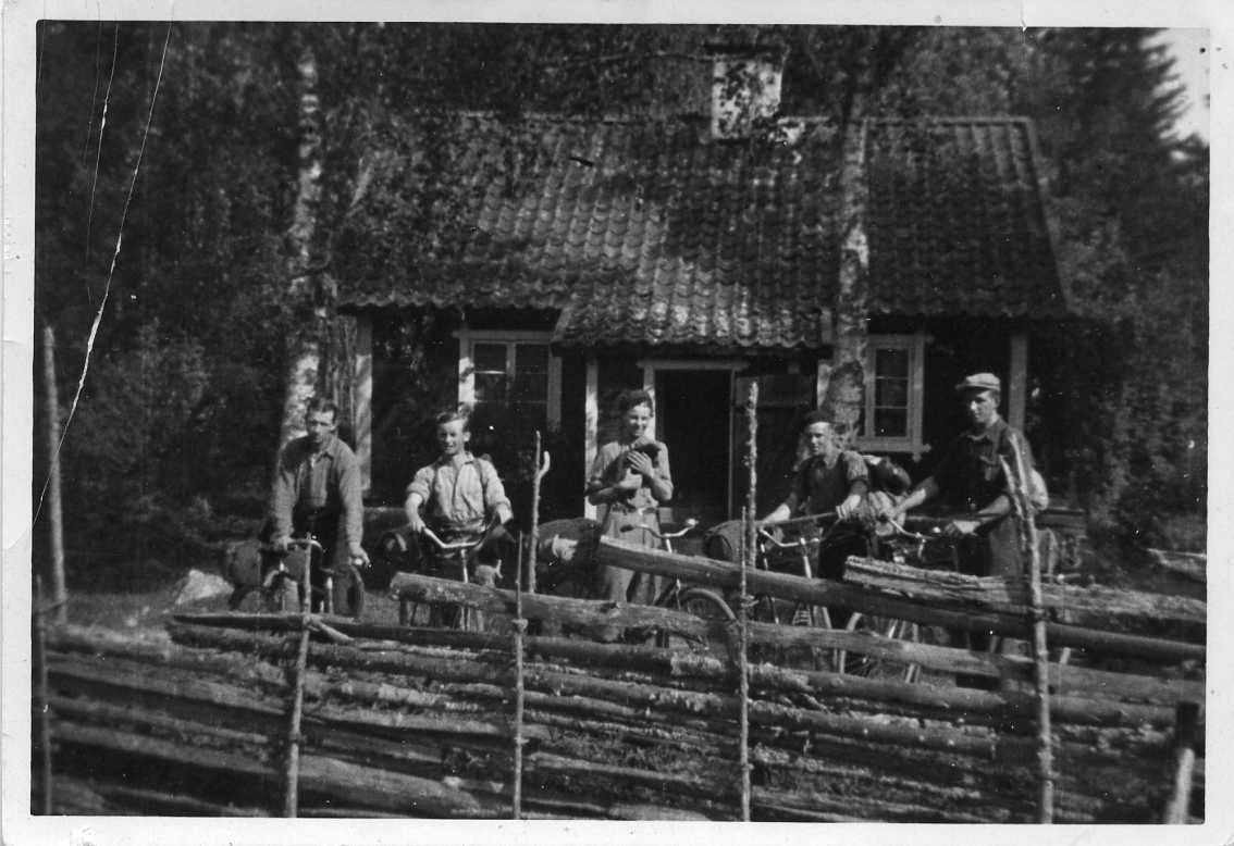 Fem unga män med ryggsäckar och cyklar står bakom en gärdesgård och framför en liten stuga. Mannen i mitten har en katt i famnen.