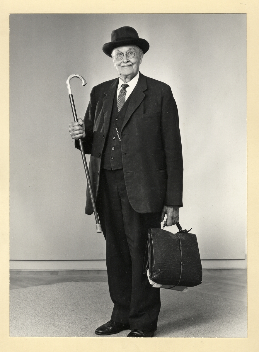 En äldre välklädd herre med glasögon och hatt, poserar vänligt för fotografen. I handen har han en käpp
med silverkrycka.