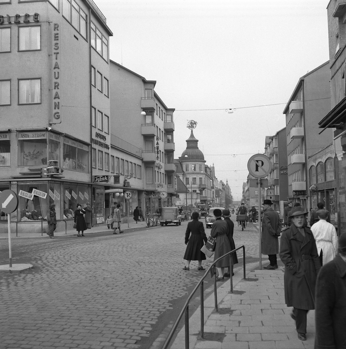 Folkliv utmed Sankt Larsgatan i höjd med Trädgårdstorget i Linköping. Året är 1962 och ännu en tid ska herrar bära hatt med självklarhet. En förändring i stadsbilden medför annars uppförandet av det så kallade Thulehuset. Byggarbetsplatsen skymtar till vänster längre ned vid gatan.