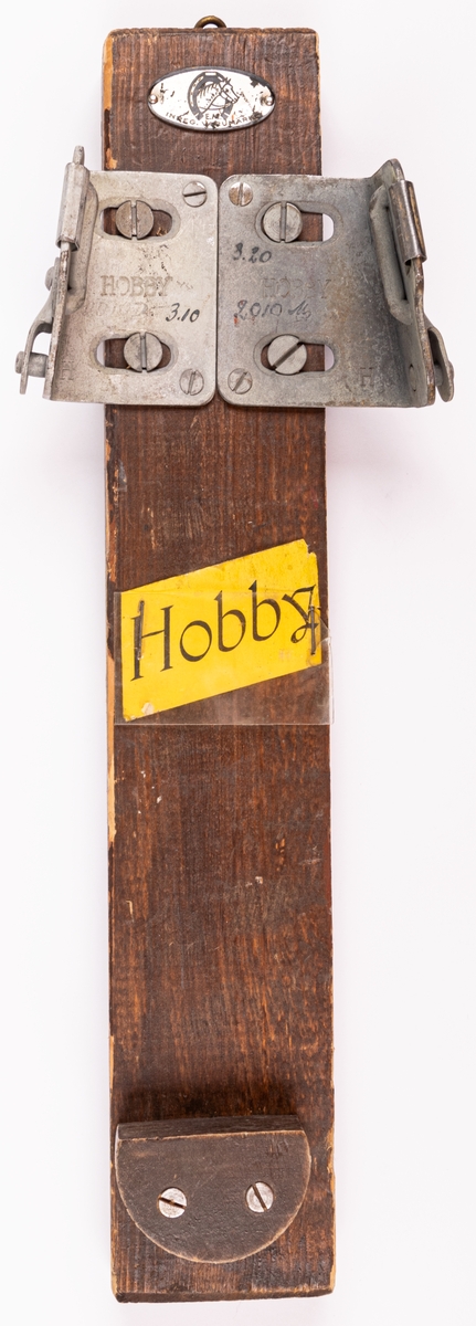 Skidbindningsprov. Vit metallegering påbrunfärgat trä. På träet en rektangulär genomskinlig plastskiva över en gul etikett med svart text "HOBBY". På metalldelarna skrift "3.20" "2010" "3.10" samt märken "HOBBY" "H". Elof Malmbergs firmamärke i metall.
