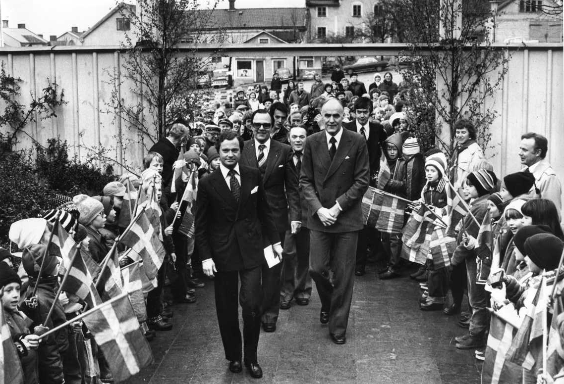 Kung Carl XVI Gustaf med flera på väg in till en samlingslokal i Gränna, torget i bakgrunden. Deras väg kantas av barn som viftar med små svenska flaggor.