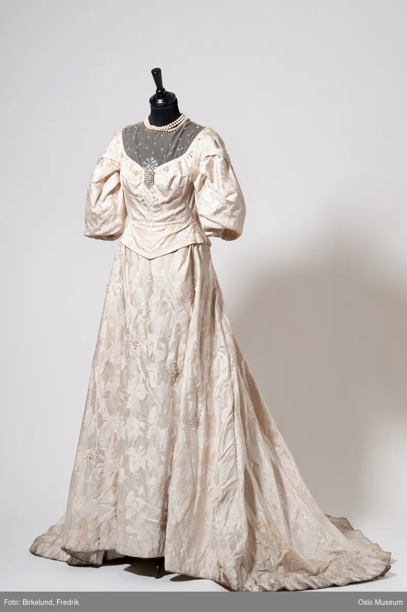 Langermet kremfarget kjole i silkebrokade med dekor av kniplinger og perler.