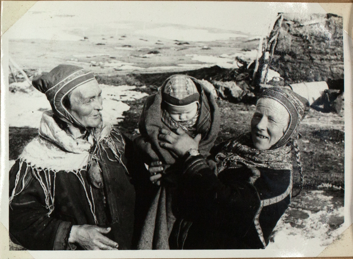 Bildet viser to eldre, samiske kvinner i tradisjonelle antrekk. De holder deres barnebarn opp. En av dem kikker i kameraet, mens den andre ser på gutten. I bakgrunnen ser man en gamme og et delvis snødekket landskap.