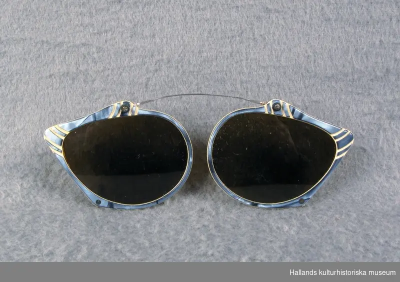 Solglasögon att sätta fast på vanliga glasögon, med blå- och vitmönstrad båge av plast. Baksidan är gulfärgad och har fyra piggar av stål att fästa med.
