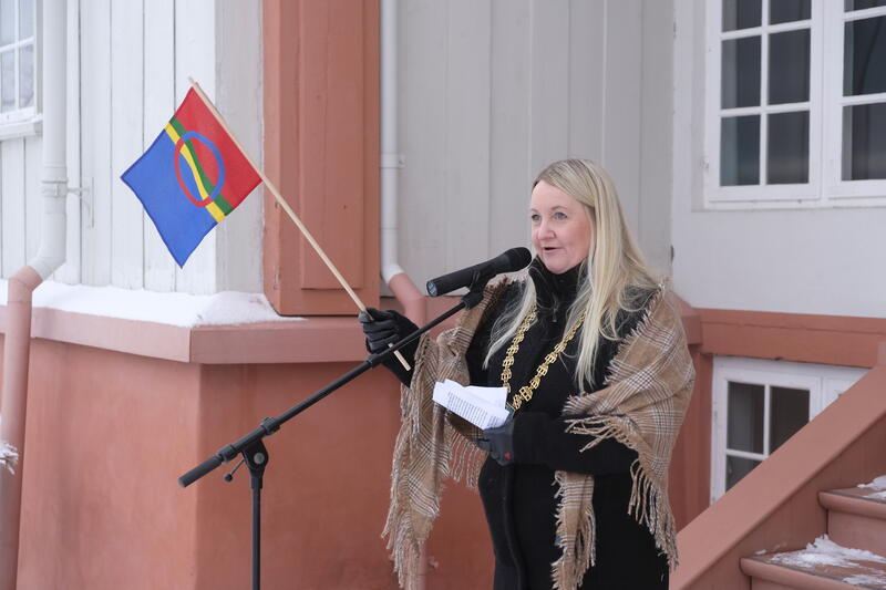 Ordfører i Eidsvoll, Hege Svendsen, halvfigur på trappa foran Eidsvollsbygningen, snakker i en mikrofon og vifter med et samisk håndflagg.