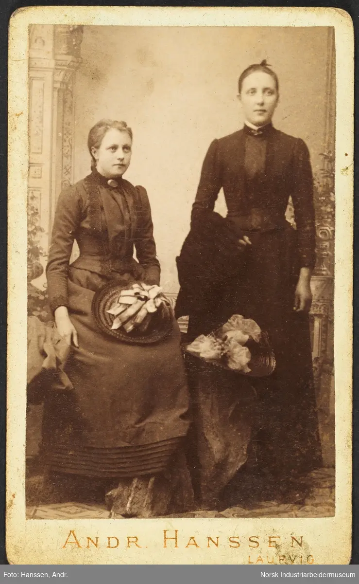 Visittkortfoto, to kvinner fotografert i studio. Begge kvinnene er kledd i pentøy, kjoler med kosettliv og sølje i halsen. Kvinnen til venstre sitter og har hatt liggende på fanget. Kvinnen til høyre står og har en jakke over den ene armen. Mellom seg har de en hatt liggende på en stein.