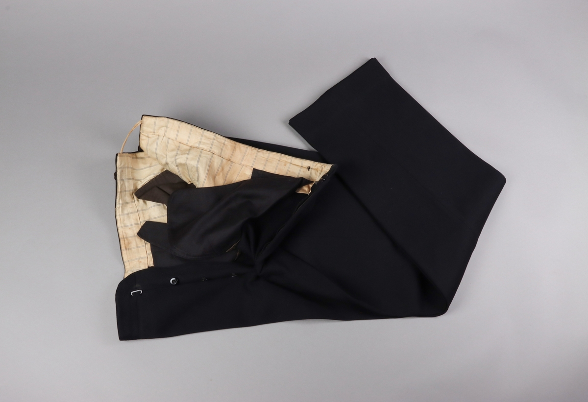 Byxor till tredelad svart kostym av kläde. Foderdelar av ljust randigt bomullstyg. Två fickor fram, en ficka bak. I ryggen bak finns ett sprund förankrat med ett tunnt band, sytt av fodertyget, som löper löst rakt över sprundet och är förankrat på varje sida. Nedanför sprundet finns en slejf med spänne för justering av passformen. Byxans bak- och sido-stycken har har iskarvningar på delarna högst upp mot linningen. Knäppgylf och knappar för hängslen.