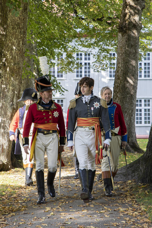 Fire menn i uniformer fra 1800-tallet går på grusgangen i alléen i Sørparken ved Eidsvollsbygningen