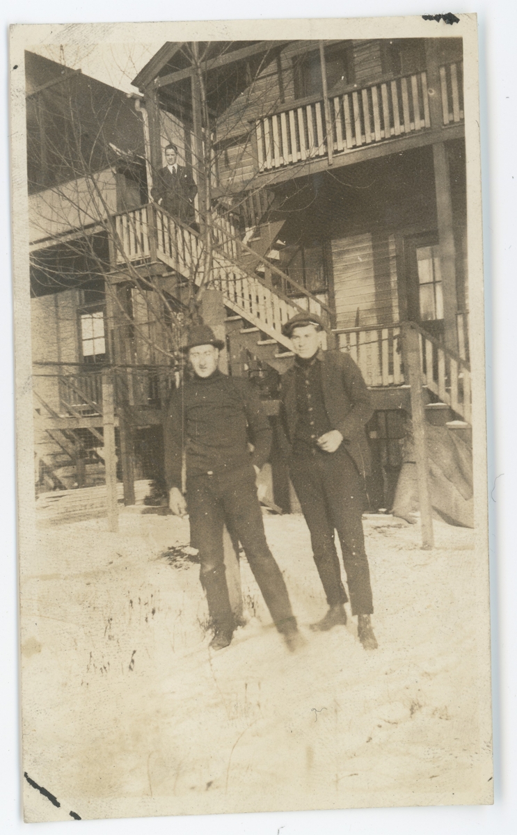To unge menn utenfor leiligheten deres i et toetasjes hus. Bjarne Fixdal står til venstre. En tredje mann står i trappa.