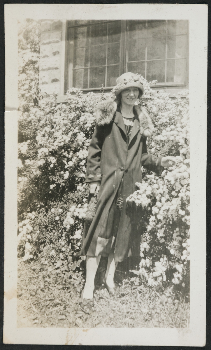 Portrett av en kvinne ved en blomsterhekk. Hun har pels og blomster i hatten.