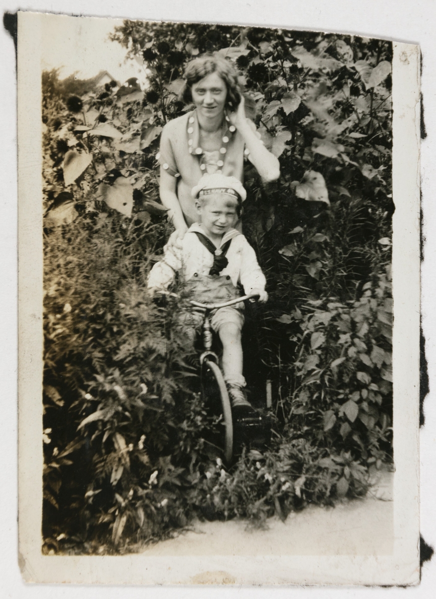 Mrs. Ragnhild Bang står bak Billy på trehjulsykkel blant solsikker og hageplanter.