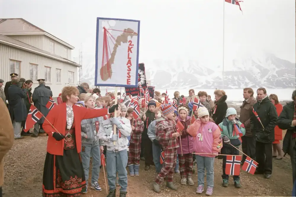 Bildet av et 17. mai tog før avmarsj. En skoleelev holder i en fane, og får hjelp av læreren iført bunad.