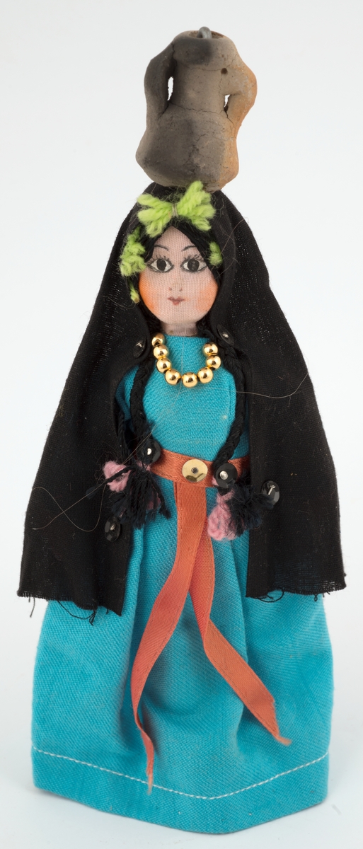 Kvinnefigur i turkis kjole, svart, langt sjal på hode; toppet av vannkrukke.